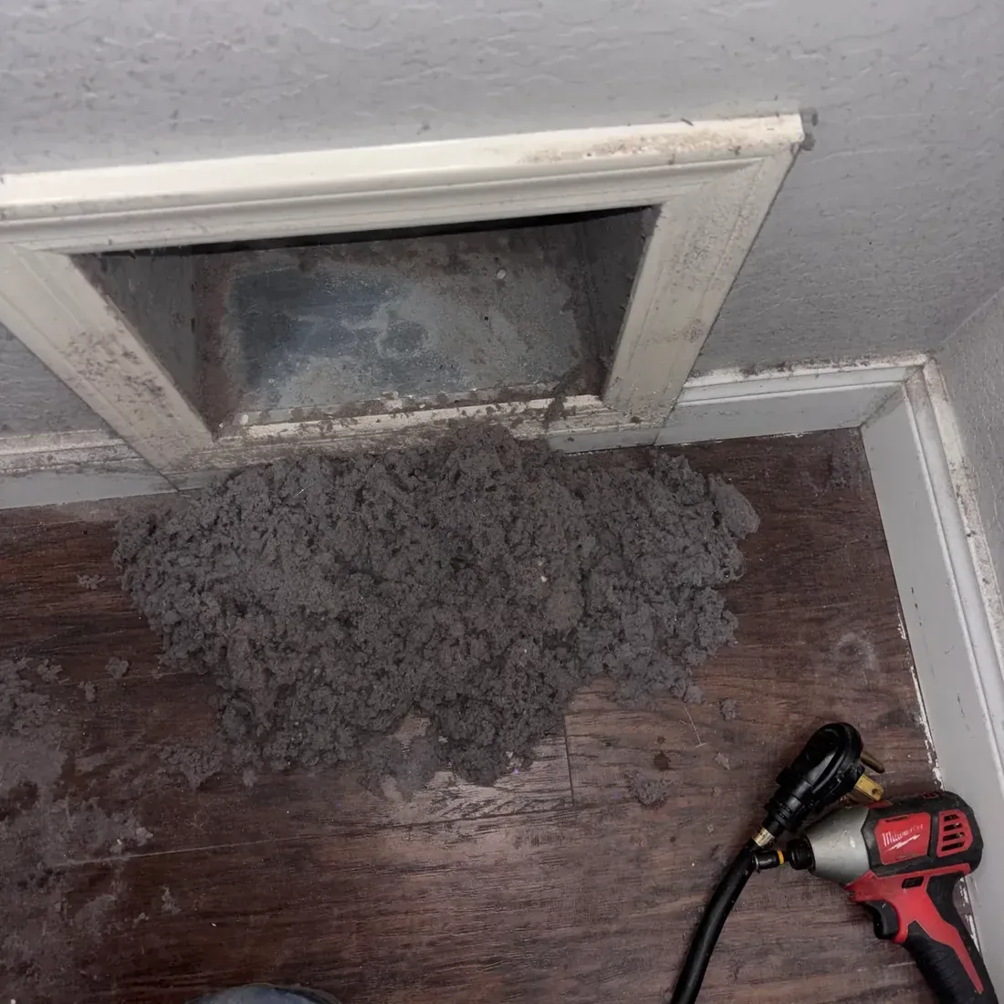 Pile of dryer lint on the floor below a wall vent opening beside a drill